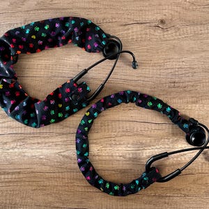 May include: Two black stethoscope covers with a rainbow paw print pattern. The covers are made of fabric and have a stretchy fit.