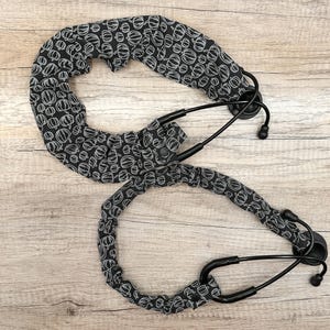 May include: Two black stethoscope covers featuring a repeating white line-art pumpkin pattern. Each cover is made of gathered fabric that wraps around the black tubing of the stethoscope, secured with small black snaps near the earpieces.