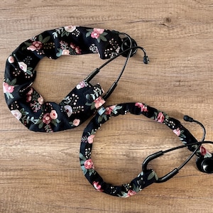 May include: Two black stethoscope covers with a floral pattern in shades of pink and green. The covers are made of fabric and have a soft, comfortable feel.