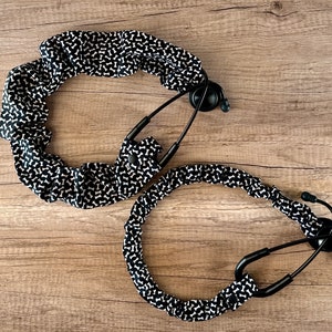 May include: Two black stethoscope covers with a white bone pattern. The covers are made of fabric and have a ruffled edge.