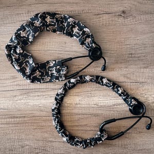 May include: Two black stethoscopes with covers. The covers are black with a repeating pattern of white skeletons. The stethoscopes are lying on a wooden surface.