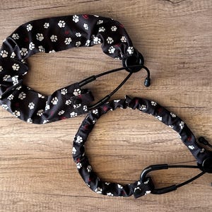 May include: Two black stethoscope covers with a white paw print pattern and small red hearts. The covers are designed to fit over the stethoscope tubing and are shown on a wooden surface.