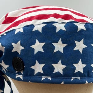 May include: A scrub cap featuring a pattern of the American flag with red and white stripes on the top and a blue field with white stars on the sides. The cap includes a black button on the side and fabric ties hanging from the back for adjustment.