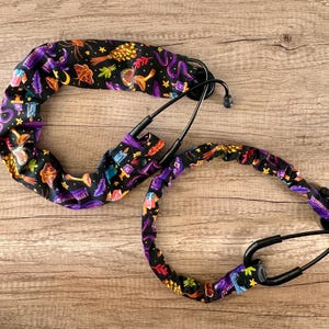 May include: Two black stethoscope lanyards with a black and purple fabric featuring a witchy pattern of brooms, cauldrons, and stars.