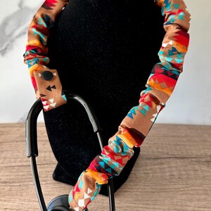 Pu&ograve; includere: Una copertura per stetoscopio in tessuto marrone con un motivo geometrico ripetuto in rosso, arancione, bianco e turchese. La copertura &egrave; arricciata lungo il tubo e include una piccola linguetta in tessuto fissata con un bottone a pressione nero.
