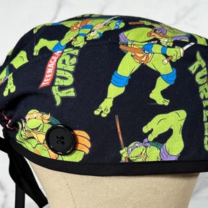 May include: A black scrub cap featuring Teenage Mutant Ninja Turtles. The cap has a repeating pattern of the turtles in action poses. The cap has a black button and black ties.