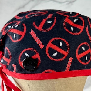 May include: Black scrub hat with a red trim and a button closure. The hat is covered in a repeating pattern of red Deadpool logos on a black background.
