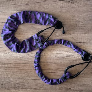 May include: Two stethoscopes with black tubing and earpieces. The stethoscope tubing is covered in a purple fabric with a repeating pattern of green bats. The stethoscopes are on a wooden surface.