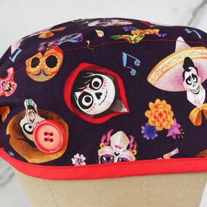 May include: A purple scrub hat with a red trim and a button. The hat features a colorful print of characters from the movie Coco, including Miguel, Hector, and the skeleton characters.