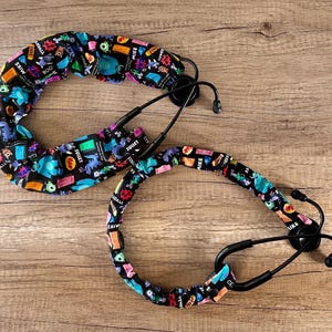 May include: Two black stethoscope covers with a colorful Monsters Inc. print. The print features characters like Mike, Sulley, and Boo. The covers are made of fabric and have a stretchy design.