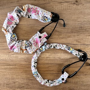 May include: A white stethoscope with a colorful patterned fabric cover featuring cartoon dogs and a confetti design. The stethoscope has a black tube and a black earpiece.
