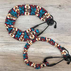 May include: A black stethoscope featuring a fabric cover with a repeating pattern of dreamcatchers and geometric shapes in shades of turquoise, red, black, and cream. The cover is secured with small black snaps along the length of the tubing.