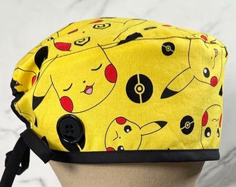 May include: Yellow scrub hat with a black border and a black button. The hat features a repeating pattern of Pikachu characters and Poké Balls.