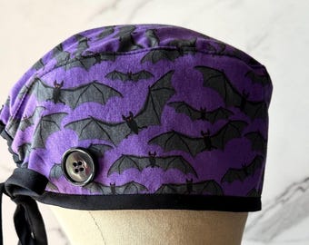 Puede incluir: Un gorro quirúrgico morado con un patrón repetido de murciélagos grises. El gorro tiene un ribete negro y un botón plateado grande. Se ata en la parte posterior con lazos negros. Diseñado para profesionales de la salud.