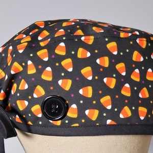May include: Black scrub hat with a candy corn print and a black button. The hat has a tie on the back.