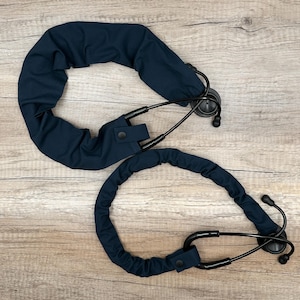 May include: Two navy blue stethoscope covers with black earpieces and chest pieces are displayed on a wooden surface. Each cover has a button closure. The covers are designed to protect the stethoscope.