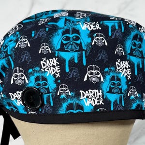 May include: A black and blue surgical scrub cap featuring a Darth Vader design. The cap has a black border and a black button. The design includes the words "Darth Vader" and "Dark Side".