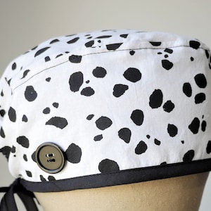 May include: A white surgical scrub cap with a black Dalmatian spot pattern. The cap has a black trim, a large black button, and black ties for securing. The fabric is a cotton blend.
