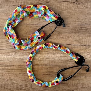 May include: Two colorful stethoscope lanyards with a gummy bear print. The lanyards are made of fabric and have a button closure. The lanyards are shown on a wooden surface.
