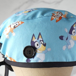 May include: Blue and black surgical scrub hat with a cartoon dog print. The hat has a black button and ties.