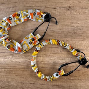 May include: Two stethoscope covers with a white background and a pattern of yellow and orange construction vehicles. The covers are made of fabric and are designed to fit around a stethoscope.