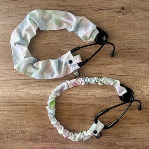 May include: Two stethoscope covers made of soft, patterned fabric. The covers are in a light blue and pink plaid pattern. The covers have a button closure and a small tag that says "Made with Love".
