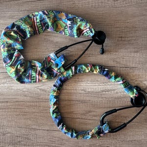 May include: Two black stethoscopes with colorful covers featuring a cartoon design. The covers are primarily blue with green, red, and yellow accents. The stethoscopes are on a wooden surface.