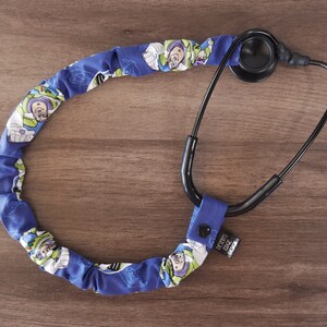 May include: A blue and green stethoscope with a fabric cover featuring a repeating pattern of the character Buzz Lightyear from the Toy Story franchise. The fabric cover is attached to the stethoscope with a black elastic band. The text "Doctor Love" is printed on the elastic band.