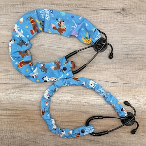 May include: Two stethoscope covers made of light blue fabric featuring a repeating pattern of various cartoon dogs, paw prints, and the text "I love my Disney dog". Each cover includes a black snap button closure to secure the fabric around the tubing.
