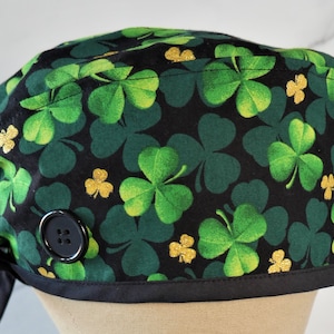 May include: Black scrub hat with a green shamrock print and gold glitter accents. The hat has a black button and ties.