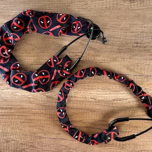 May include: Two stethoscope covers with a black background and a repeating red and white Deadpool logo pattern. The covers are designed to fit over the stethoscope tubing and earpieces. The stethoscope is black.