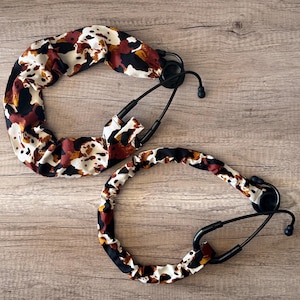 May include: Two stethoscopes with black tubing and earpieces. The stethoscope tubing covers have a cow print design in shades of brown, black, and white. The stethoscopes are lying on a wooden surface.