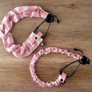 May include: Two pink and white gingham stethoscope covers. One cover is a long tube with a snap closure. The other cover is a shorter tube with a snap closure. Both covers are made of soft fabric.