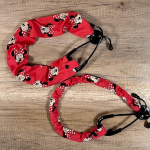 May include: Two red stethoscope covers with a Minnie Mouse pattern. The covers are made of fabric and feature a repeating pattern of Minnie Mouse's face and bow. The stethoscope's black earpieces and chest piece are visible.