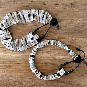May include: Two black stethoscope holders with a mummy pattern. The holders are made of fabric and have a white background with black mummy bandages and eyes.