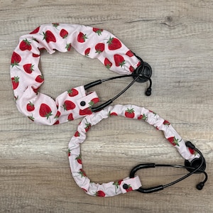 May include: Two stethoscopes with black tubing and earpieces. The tubing is covered in a light pink fabric with a pattern of red strawberries and white polka dots. The fabric covers have a button closure.