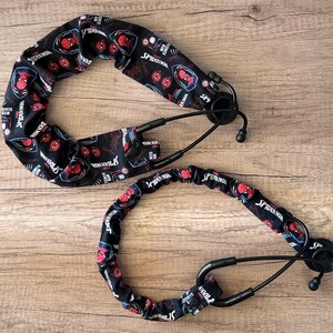 May include: Two black stethoscopes with Spider-Man themed covers. The covers are black with red Spider-Man faces and the words "Spider-Man" printed on them. The stethoscopes are on a light brown wooden surface.