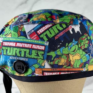 May include: A black and blue scrub hat with a Teenage Mutant Ninja Turtles comic book print. The hat has a black button and a black tie.