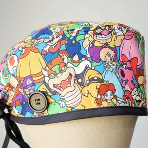 May include: A colorful surgical scrub hat with a black trim and a button. The hat features a pattern of various characters from the Super Mario Bros. video game franchise, including Mario, Luigi, Princess Peach, Bowser, and Toad.