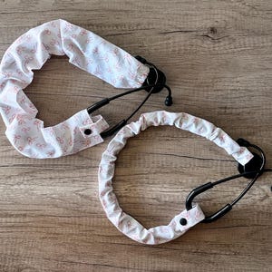 May include: Two stethoscopes with covers. The covers are white with a pink bow pattern. The stethoscopes are black and rest on a wooden surface.