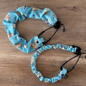 May include: A blue stethoscope with a patterned fabric cover featuring cartoon dogs. The fabric cover is elastic and has a pocket for the stethoscope's earpieces. The fabric cover is blue with white and brown cartoon dogs.