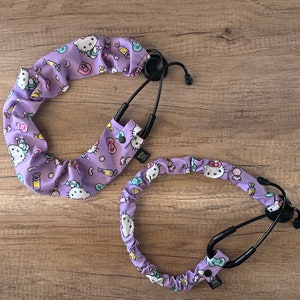 May include: Two purple stethoscope holders with a Hello Kitty print. One is a long, thin holder with a black stethoscope attached. The other is a shorter, thicker holder with a black stethoscope attached.