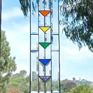 May include: A rectangular stained glass window panel with a black metal frame. The panel features a series of colorful inverted triangles in red, orange, yellow, green, blue, and purple. The background shows a bright blue sky and green trees.