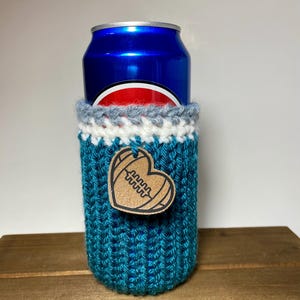 Op de afbeelding: Een turquoise gebreide blikjeshoes met een witte, grijze en turquoise rand. Een houten hartvormig label met een voetbalontwerp is aan de hoes bevestigd. De hoes bevat een blauw blikje.