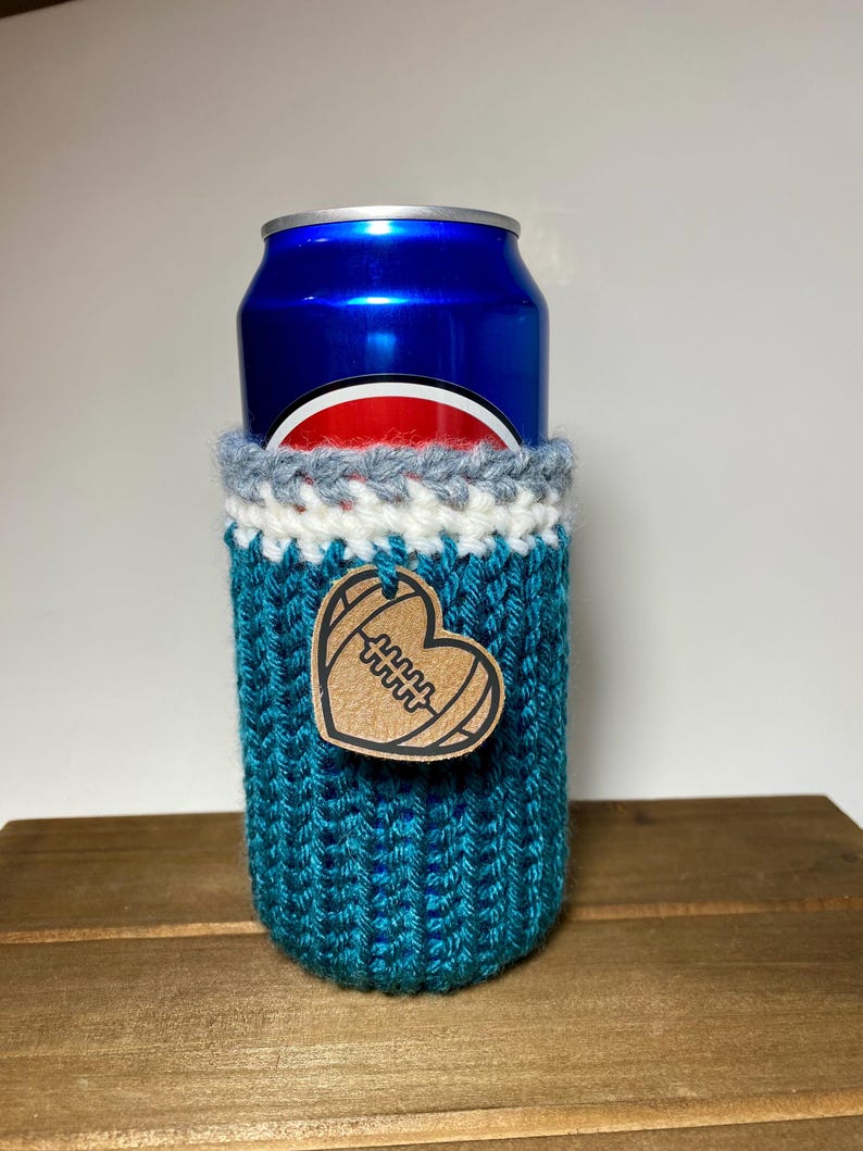 Op de afbeelding: Een turquoise gebreide blikjeshoes met een witte, grijze en turquoise rand. Een houten hartvormig label met een voetbalontwerp is aan de hoes bevestigd. De hoes bevat een blauw blikje.