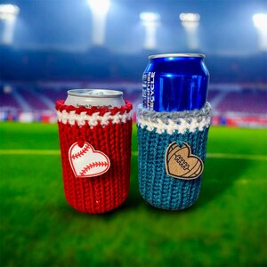 Op de afbeelding: Twee gebreide blikjeskoelers, &eacute;&eacute;n rood met een honkbalhart, de andere blauwgroen met een voetbalhart. De koelers staan op een groen veld met een stadionachtergrond. De rode koeler heeft de tekst "Supports Digestive Health".
