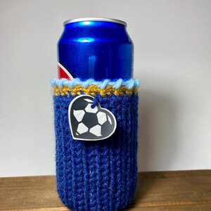 Op de afbeelding: Een blauwe gebreide blikjeshoes met een hartvormig voetbaltag. De hoes is om een blauw blikje gewikkeld. De bovenkant van de hoes heeft een gele en blauwe rand. De hoes staat op een houten oppervlak.