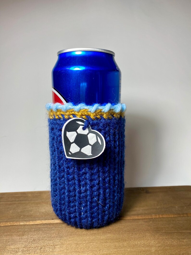 Op de afbeelding: Een blauwe gebreide blikjeshoes met een hartvormig voetbaltag. De hoes is om een blauw blikje gewikkeld. De bovenkant van de hoes heeft een gele en blauwe rand. De hoes staat op een houten oppervlak.
