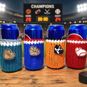 May include: Four blue beverage cans in knitted cozies with sports-themed heart-shaped tags. The cozies are teal, blue, orange, and red, with tags featuring a football, basketball, hockey, and baseball. A football, baseball, and basketball are also visible.