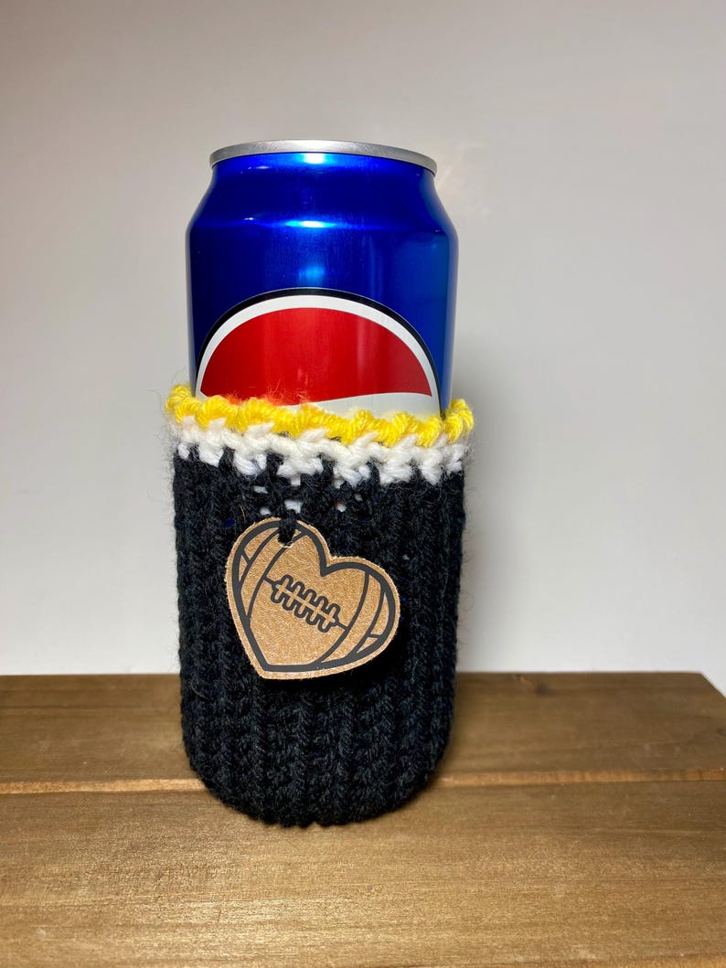Op de afbeelding: Een blauw frisdrankblikje in een zwarte gebreide bekerhouder met gele en witte rand. Een hartvormig label met een voetbalontwerp hangt aan de bekerhouder. De bekerhouder is ontworpen om dranken koud te houden.